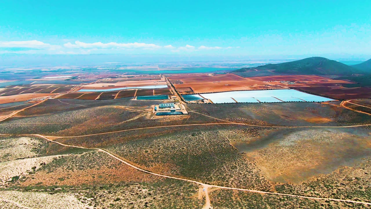 San Quintin BC Mexico aerial frame showing a broad dry plain with intersecting access roads, clustered small structures near the ponds, distant cultivated-looking rectangles, and no visible people.