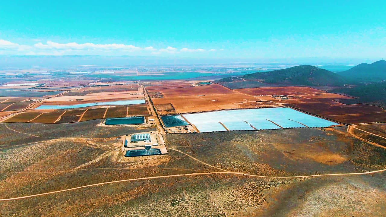 High aerial view over San Quintin BC Mexico showing dry scrubland in front, a solar panel site, rectangular water reservoirs, reddish fields, distant hills, clear sky, and no visible people.