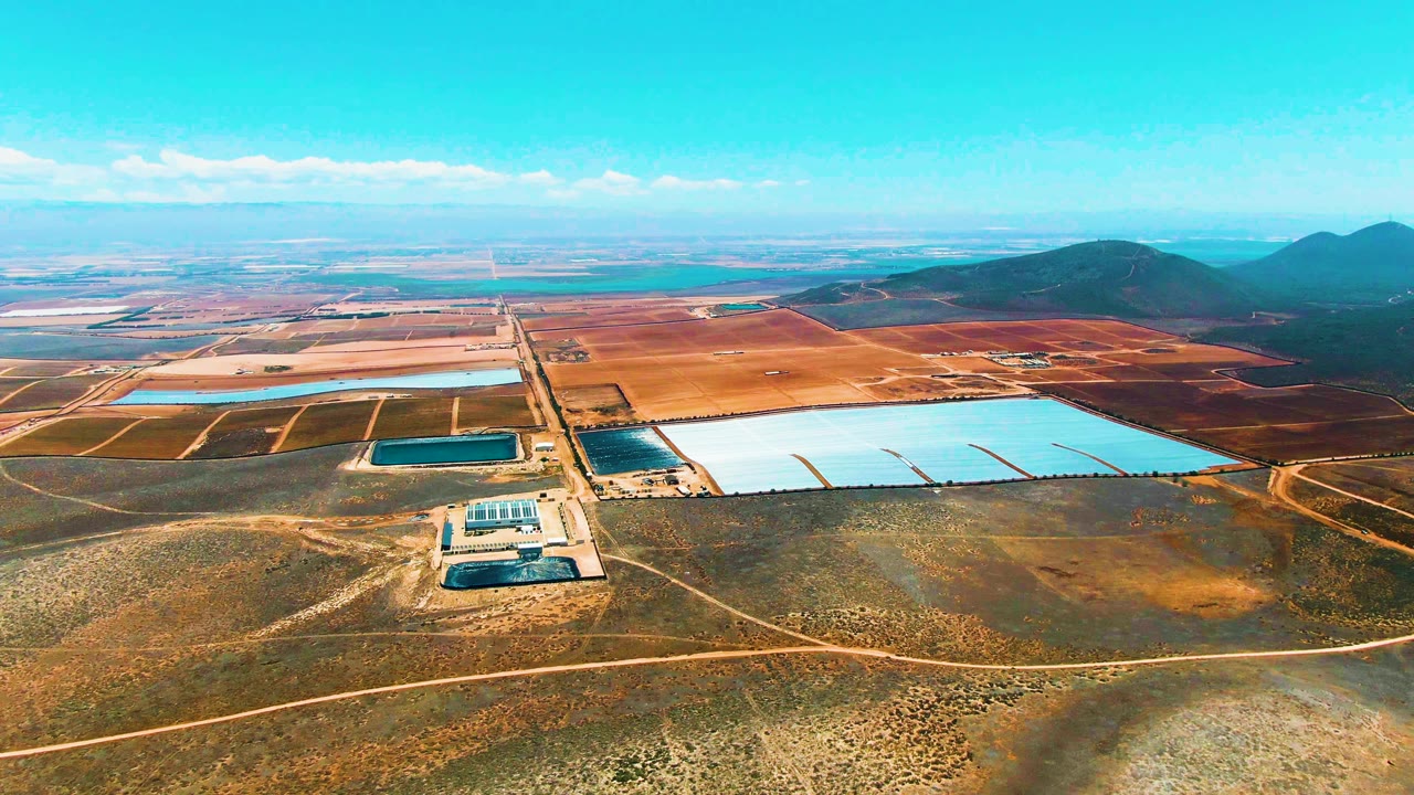 Aerial frame from San Quintin BC Mexico with pale reservoir basins crossing the midground, a small solar panel array below them, brown agricultural blocks, dark hills, blue sky, and no visible people.