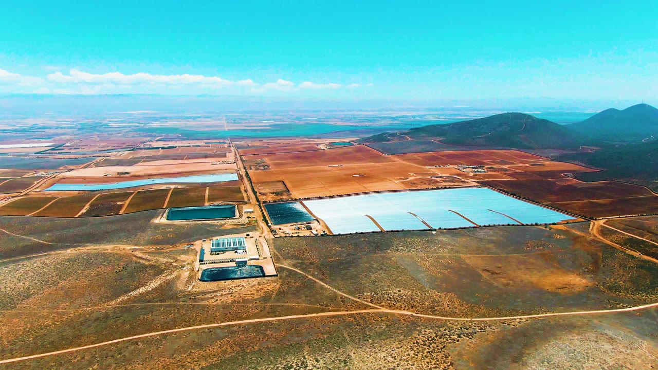 Wide San Quintin BC Mexico aerial showing the reservoirs stretching across the right midground, a solar installation near the lower center, branching dirt tracks, arid foreground, distant fields, and no visible people.