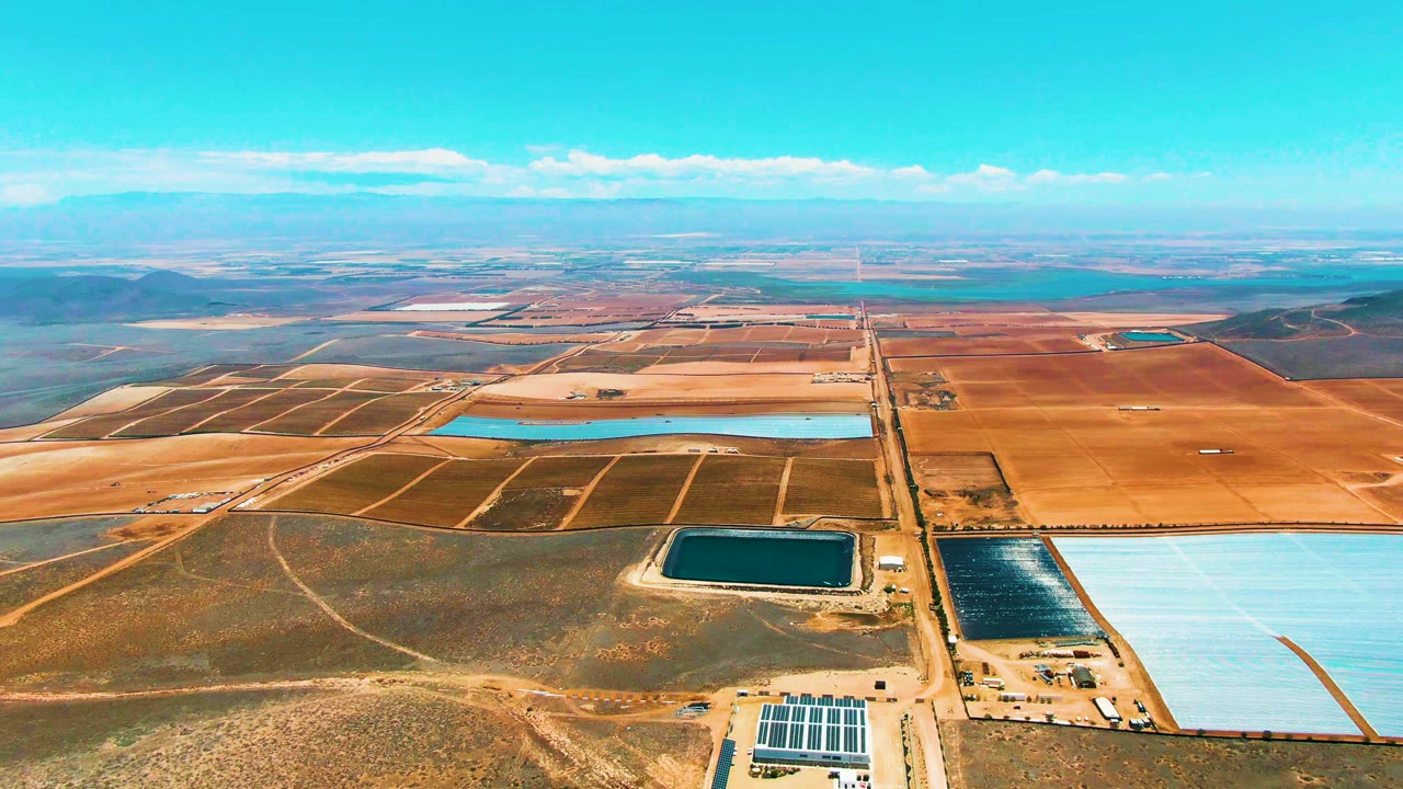 San Quintin BC Mexico aerial view with a central road dividing dry fields, turquoise and dark blue reservoirs, faint field boundaries, distant terrain, scattered clouds, and no visible people.