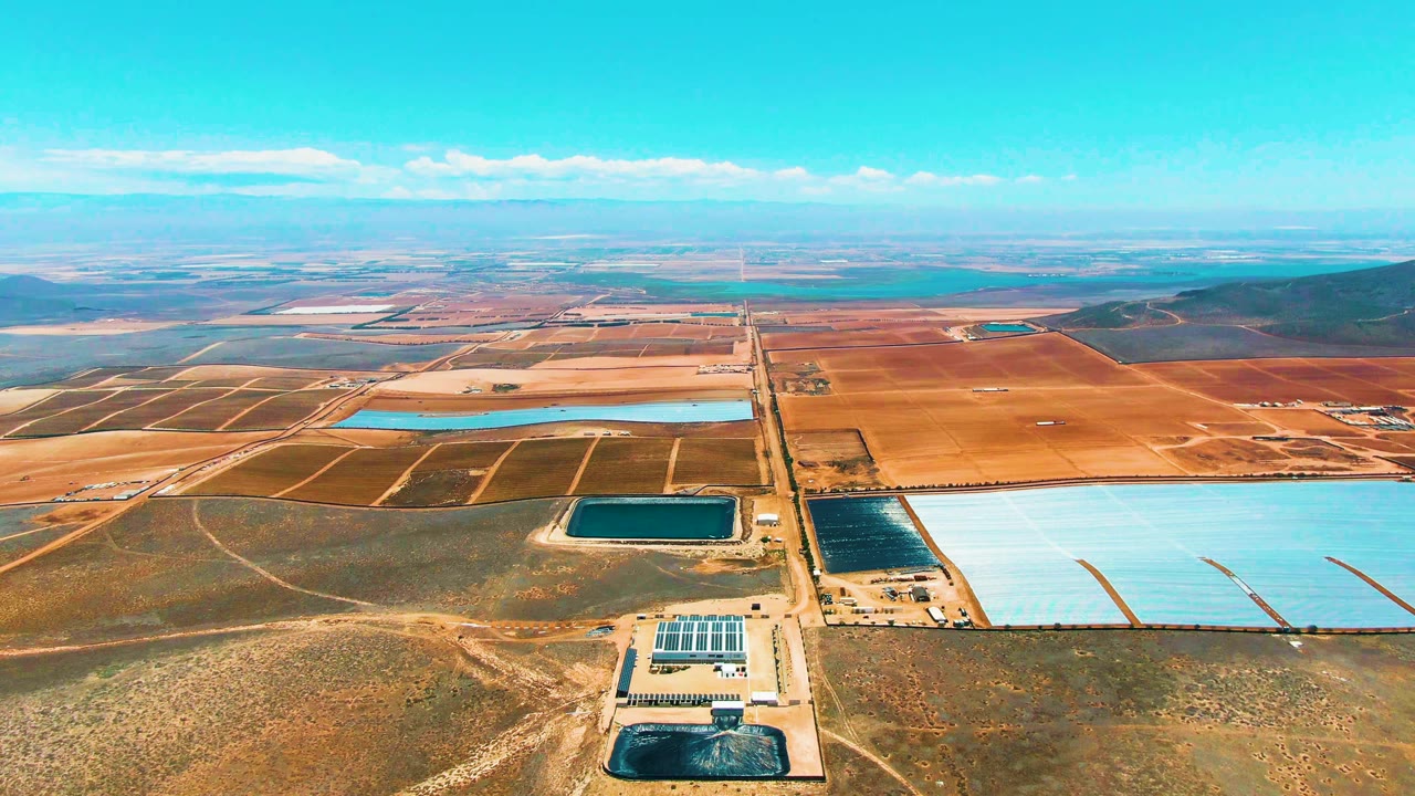 Aerial frame over San Quintin, Mexico showing blue reservoirs, a solar facility near the lower edge, dry brown terrain, distant fields, low hills, and no visible people.