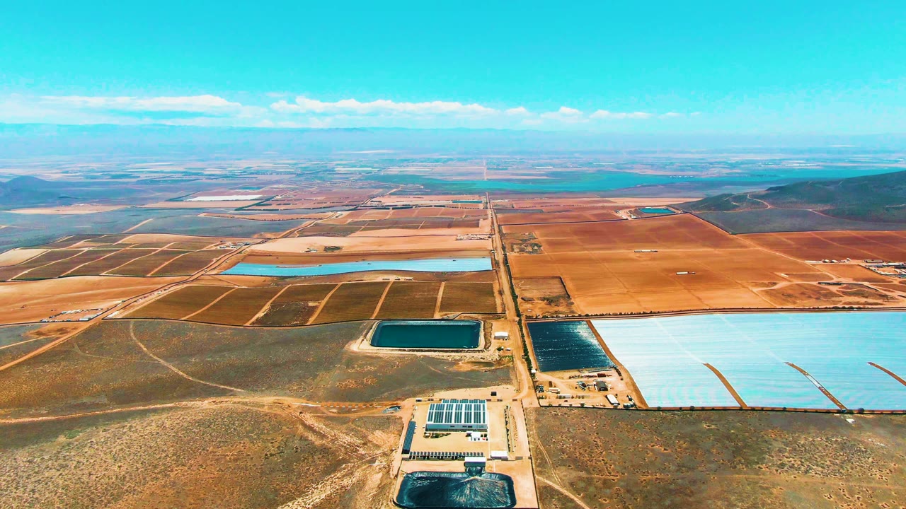 Second aerial frame over San Quintin, Mexico with the solar facility near the lower center, dark and pale blue reservoirs to the right, a straight road through brown fields, and no visible people.