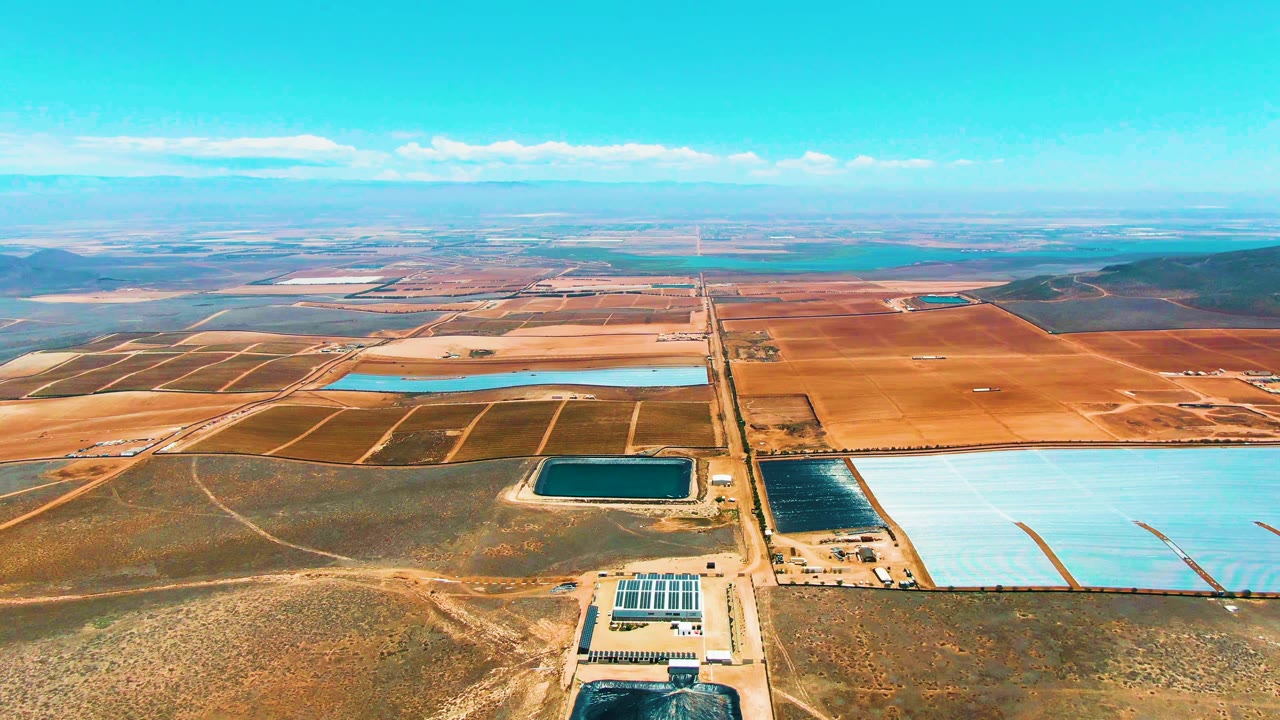 Third frame from the San Quintin aerial shows large pale reservoirs on the right, darker basins near the road, a compact solar array at the bottom, open brown land, and no visible people.