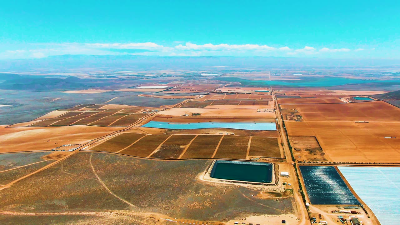 High aerial view over San Quintin, Mexico with turquoise and dark green evaporation ponds, beige field roads, distant low hills, and no visible people.