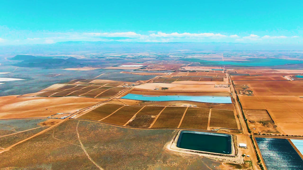 High aerial view over San Quintin BC, Mexico farmland with brown field blocks, blue reservoirs, distant low hills, and no visible people at the opening moment.