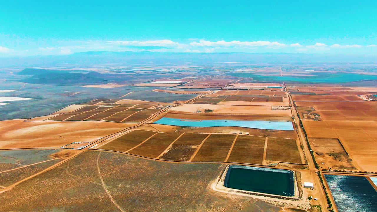 Aerial frame over San Quintin BC, Mexico showing rectangular field grids, a bright blue reservoir across the center, darker water basins near the bottom edge, and no visible people.