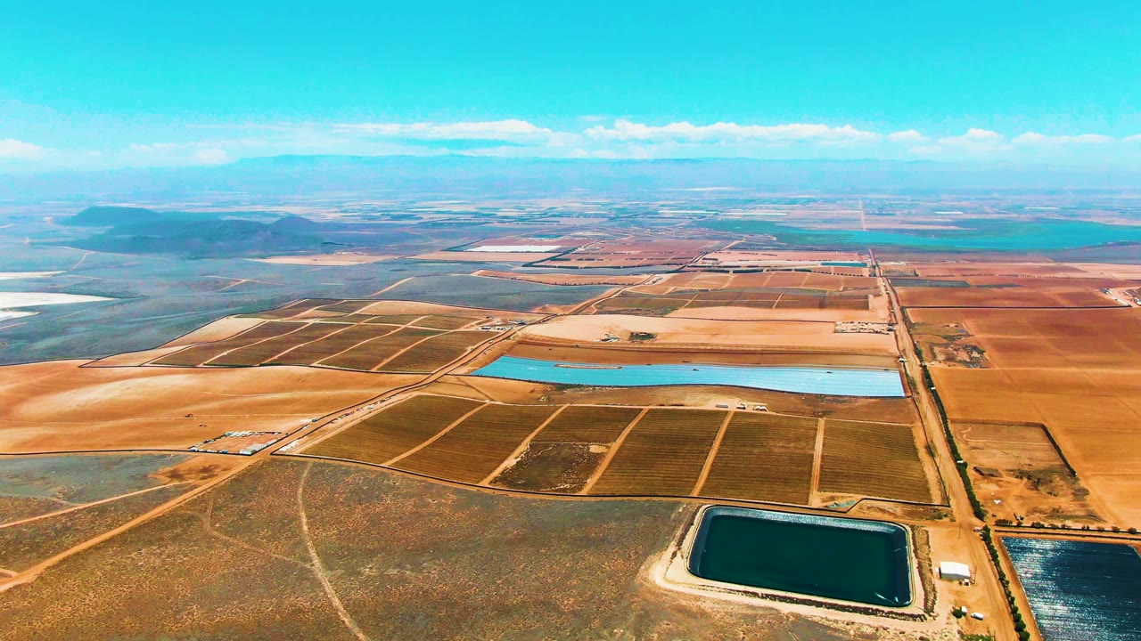 High drone view in San Quintin BC, Mexico with dirt roads dividing brown and green fields, a long blue reservoir in the midground, distant flatlands, and no visible people.