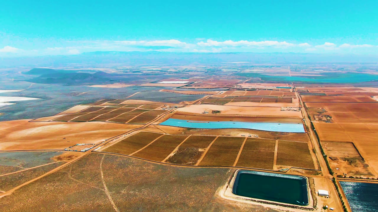 Aerial view across San Quintin BC, Mexico fields with a turquoise reservoir spanning the center, darker reservoir water near the lower edge, low hills in the distance, and no visible people.