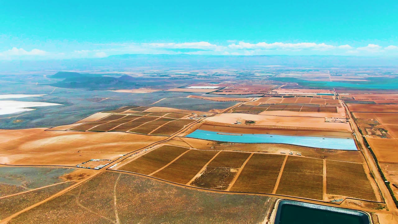 Second aerial frame over San Quintin BC, Mexico, with a long turquoise reservoir centered among rectangular field divisions, a straight road at right, and no visible people.