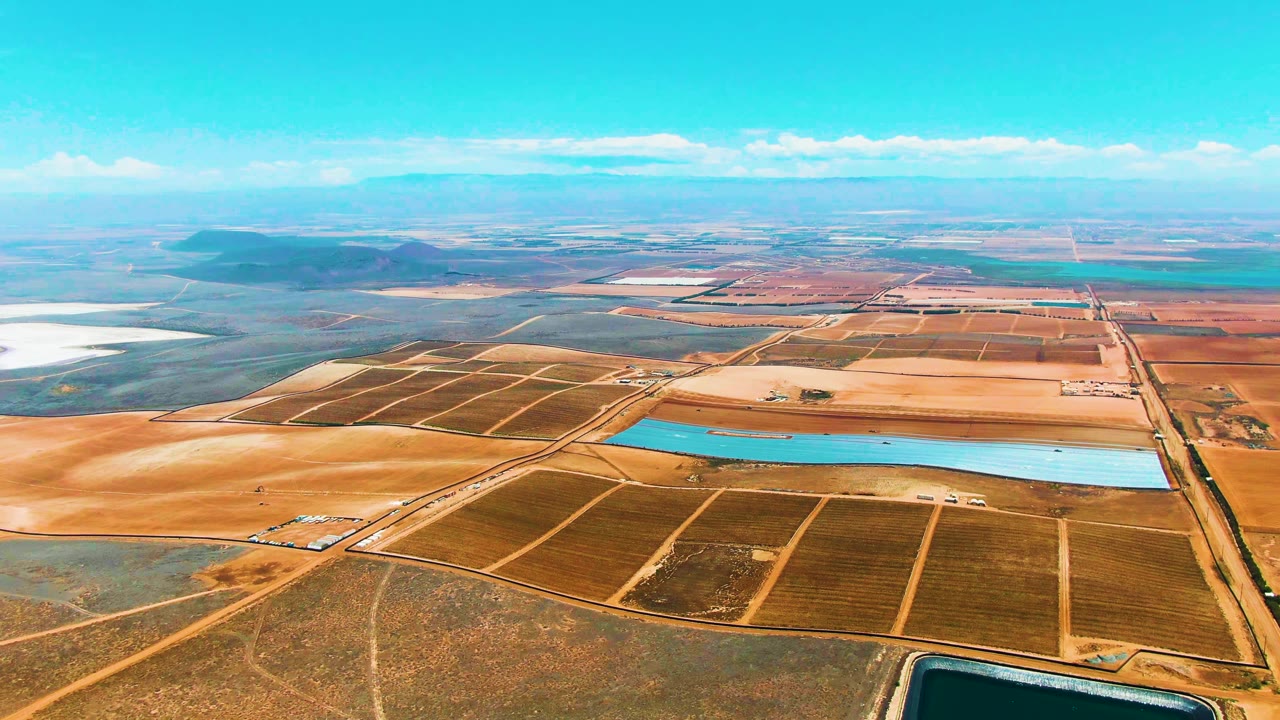 Third aerial frame over San Quintin BC, Mexico, showing the reservoir stretched across the midground, darker planted fields below it, pale dry plots beyond it, and no visible people.