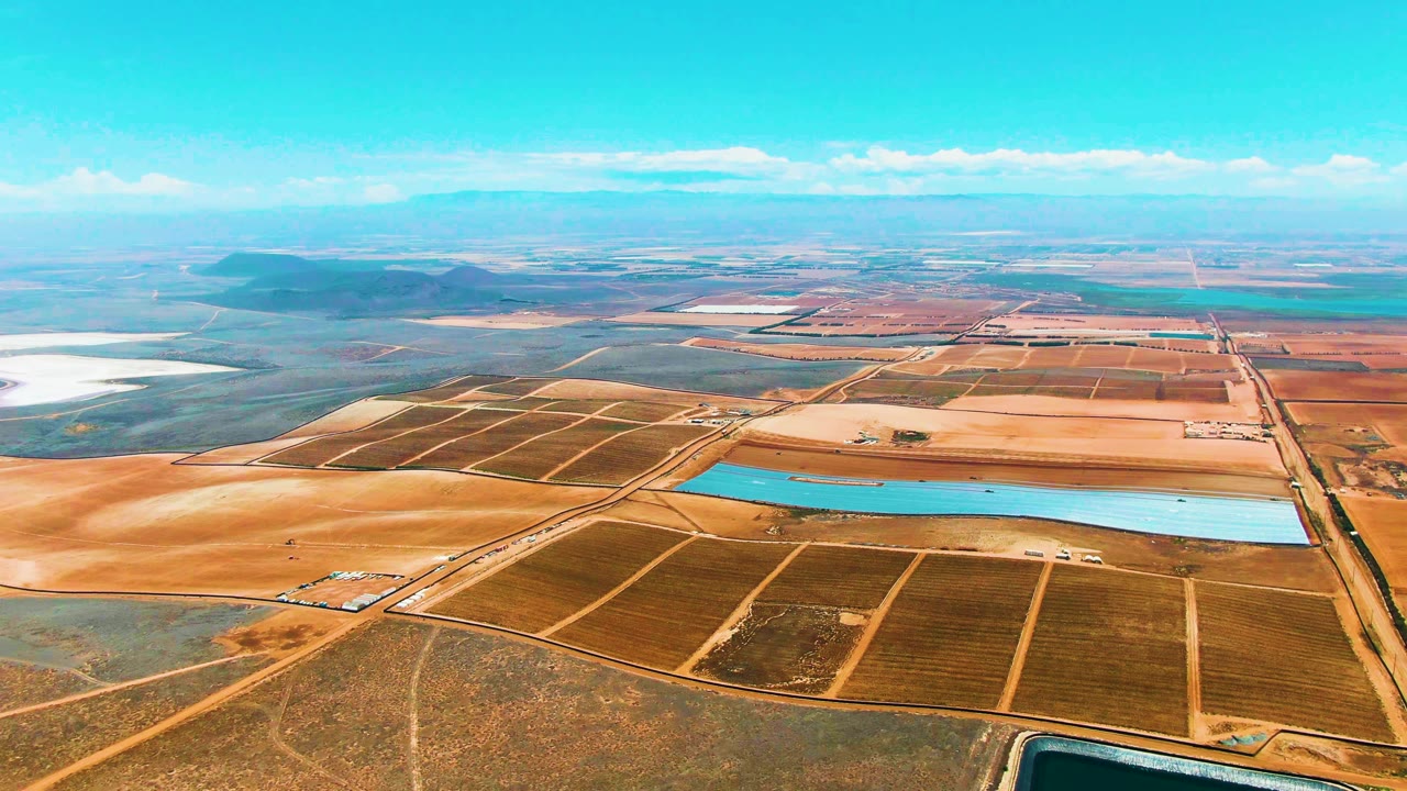 Aerial view across arid farmland with a large blue pool and horizon.