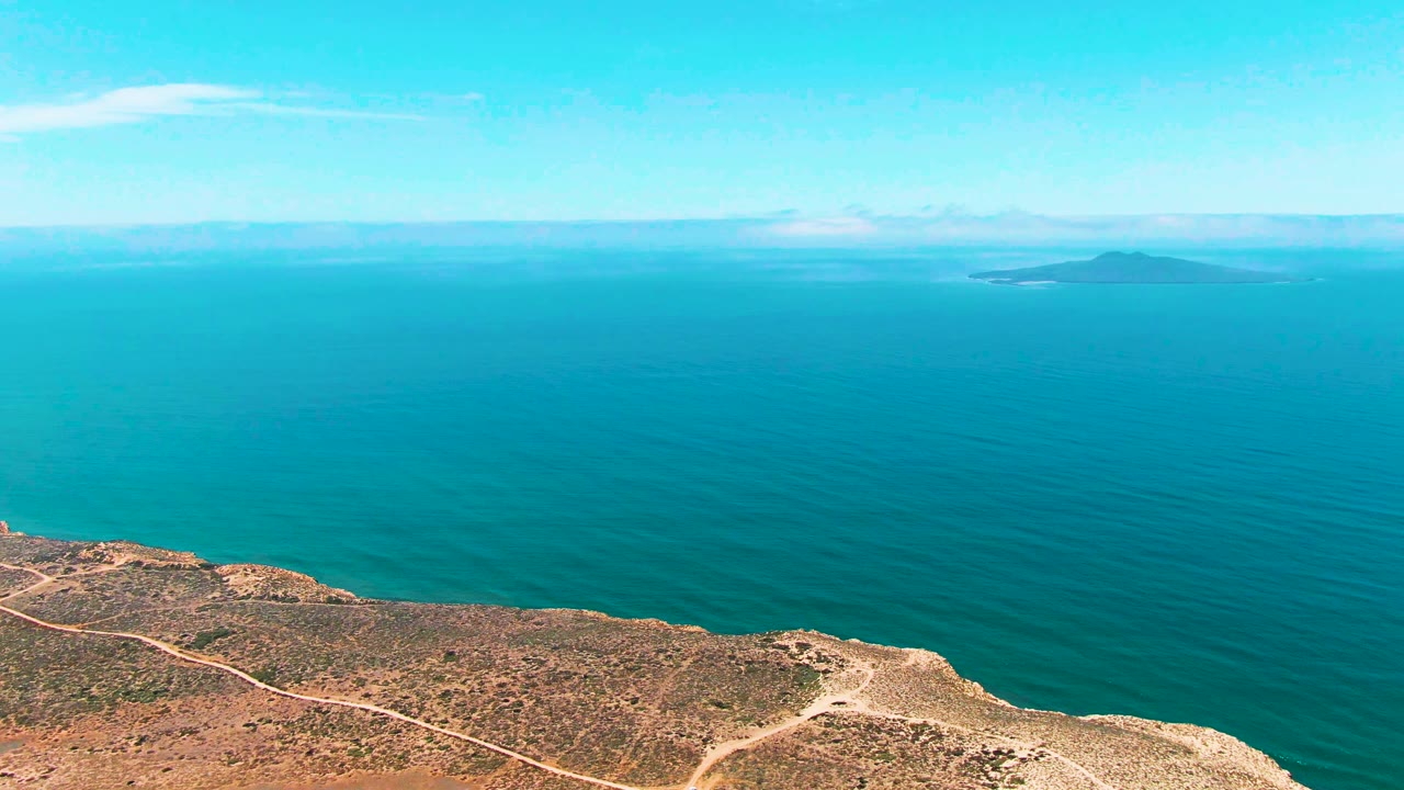 Rocky cliff above turquoise ocean with distant island on the horizon.
