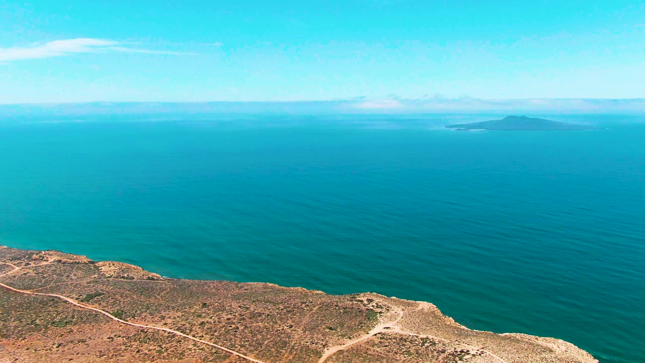 One second into the San Quintin BC Mexico clip, dry cliff terrain and pale dirt tracks sit below calm blue-green ocean, with the island still visible on the horizon and no visible people.