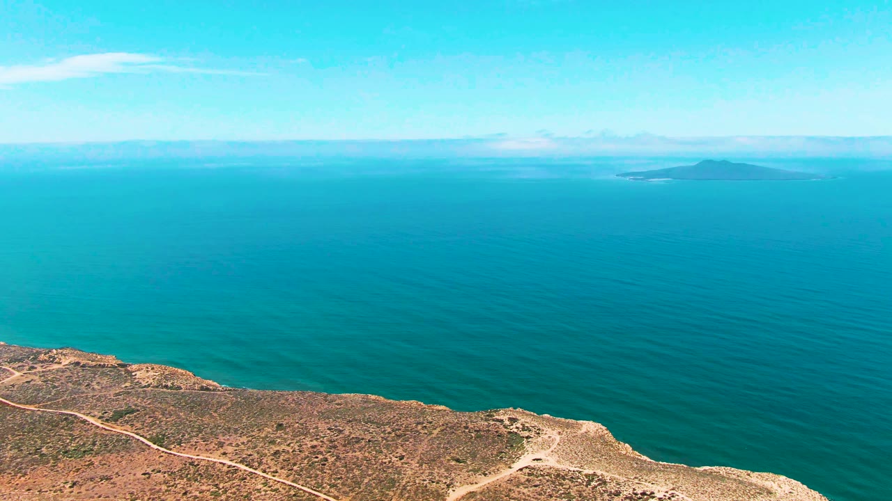At two seconds, the San Quintin BC Mexico cliff remains low in frame while turquoise sea fills most of the view, a low island sits slightly right of center on the horizon, and no visible people appear.