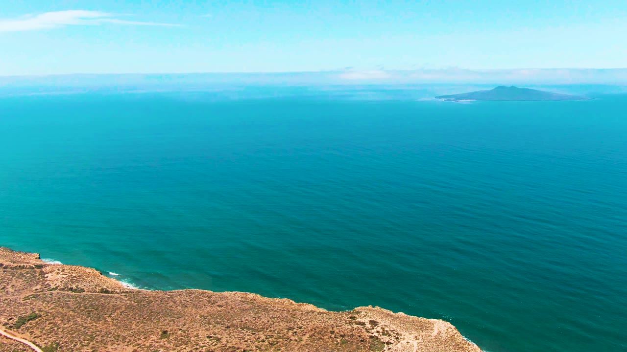 A wide elevated view in San Quintin BC Mexico shows turquoise ocean, a dry tan cliff edge at the bottom, distant low islands on the horizon, and no visible people.