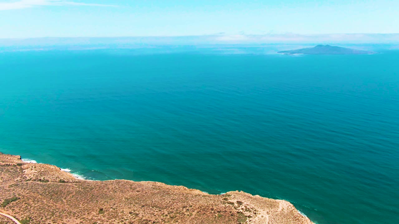 At 0:02, the aerial view shows the San Quintin BC Mexico cliff running along the bottom, deep blue-green ocean stretching outward, faint islands near the horizon, and no visible people.