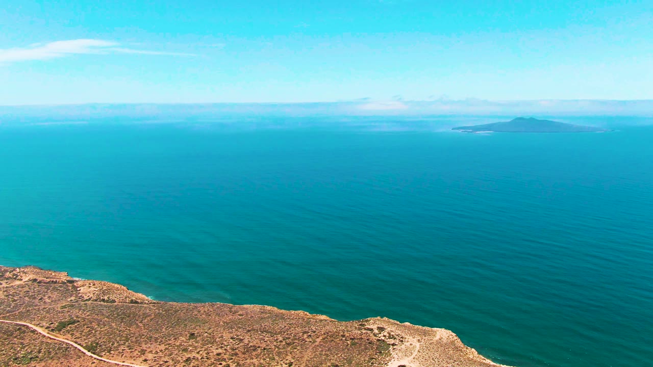 Aerial frame from San Quintin BC Mexico with the dry cliff slightly more centered along the bottom, rippled blue water across the middle, a far island, and no visible people.
