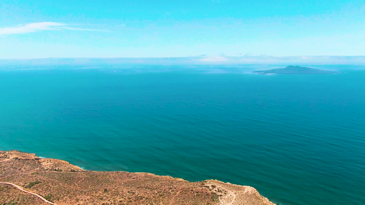 San Quintin BC Mexico coastline seen from above, with a narrow rocky shore at the bottom, teal ocean stretching to a small distant island, pale clouds, and no visible people.