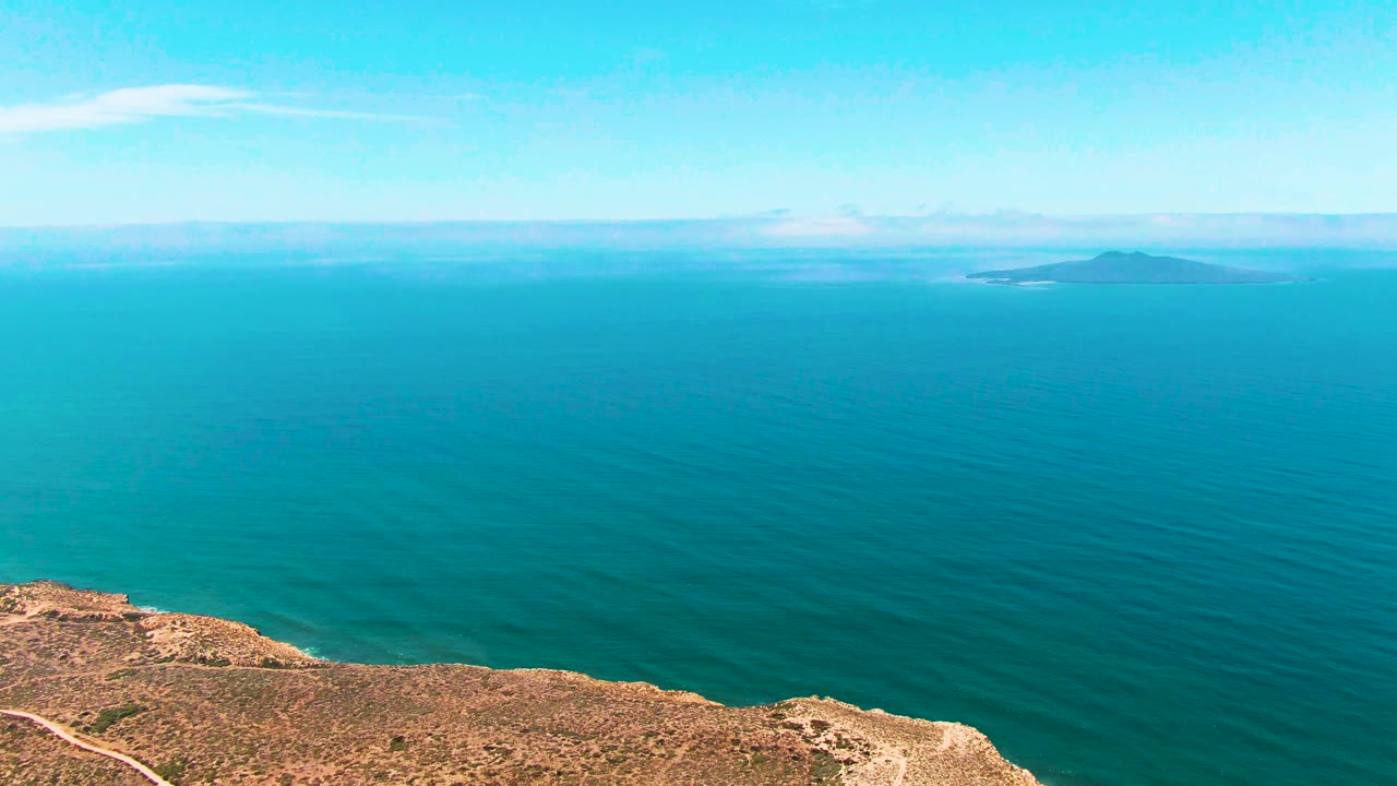 Aerial San Quintin BC Mexico view with the cliff entering from the lower left, dark and light bands in the ocean surface, a distant island near the horizon, and no visible people.