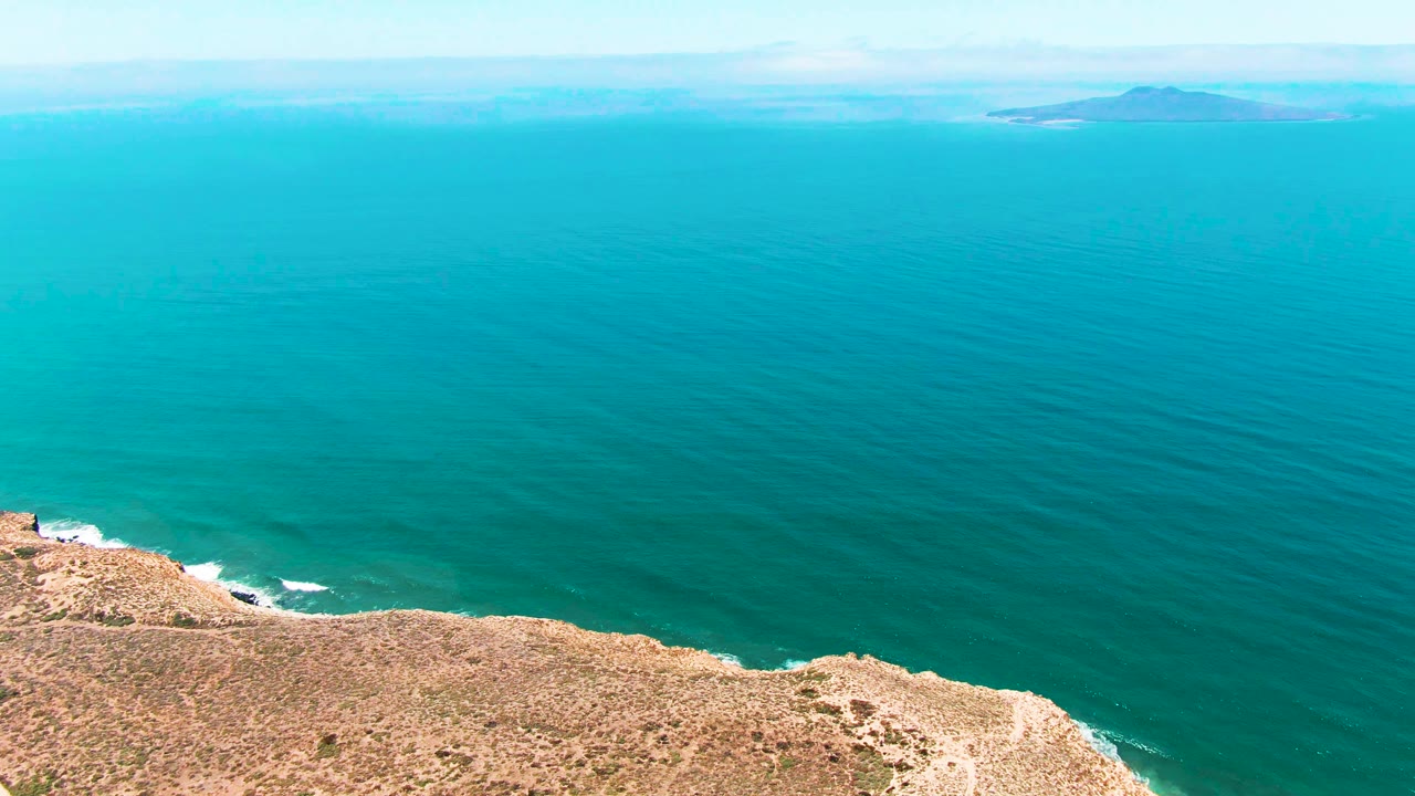 Aerial view in San Quintin BC Mexico with a rocky cliff along the lower edge, turquoise ocean across the frame, faint distant islands on the horizon, and no visible people.