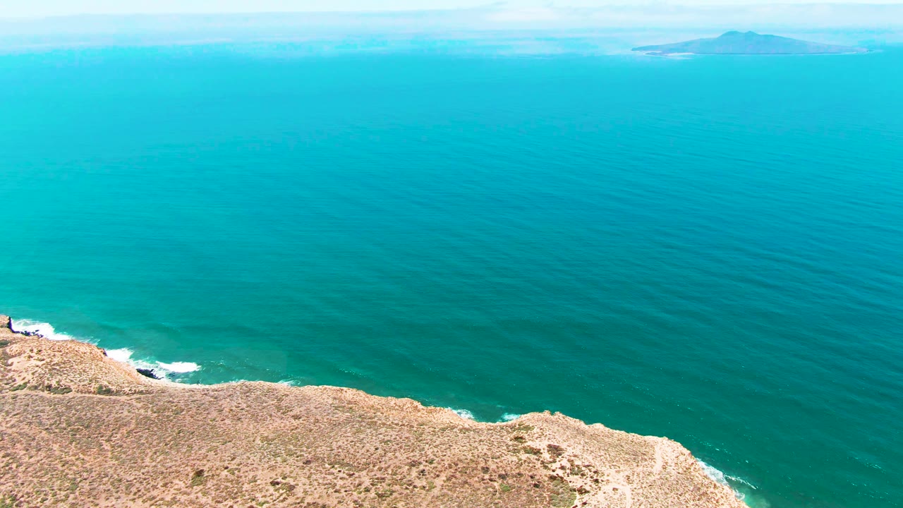 Aerial coastal frame from San Quintin BC Mexico showing the cliff line low in frame, turquoise ocean texture, pale sky at the horizon, distant island forms, and no visible people.