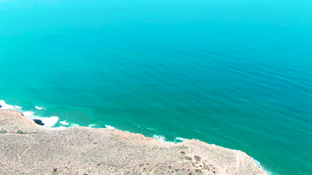 Opening aerial view of turquoise water breaking into white foam along the rocky San Quintin BC Mexico shoreline, with no visible people.