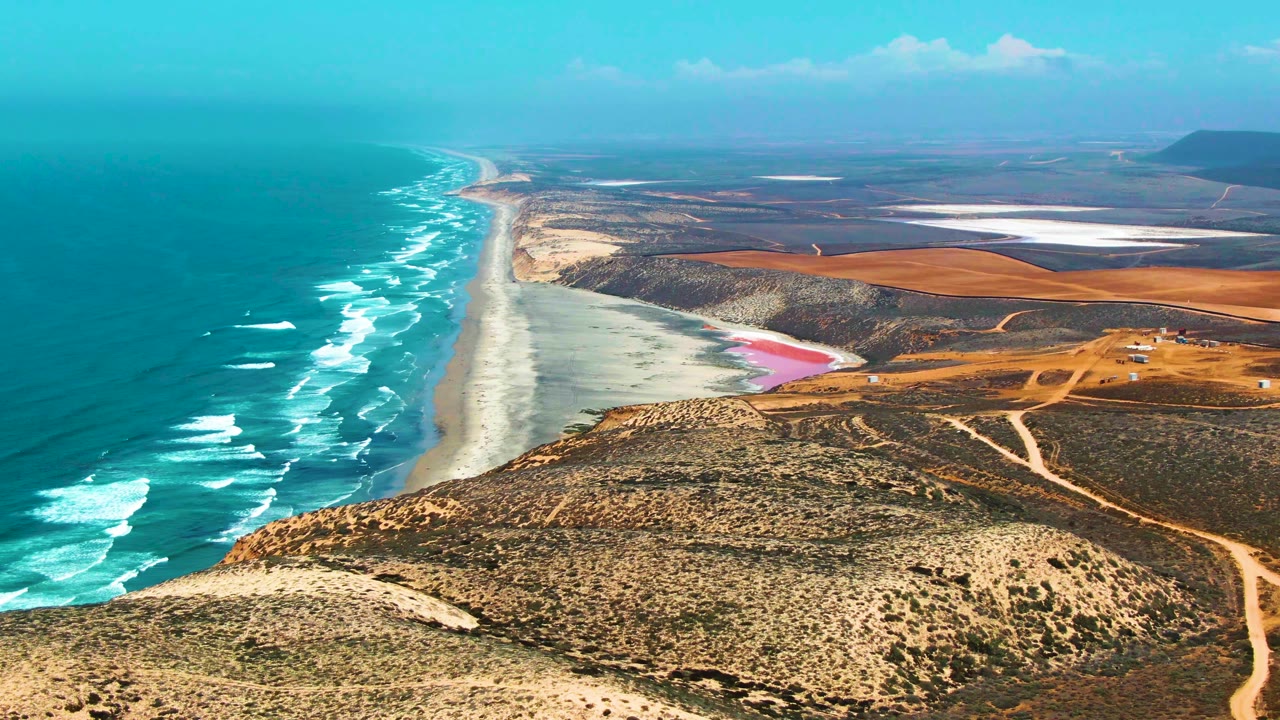 Opening aerial frame over San Quintin BC Mexico, with turquoise ocean and white surf on the left, scrubby coastal hills in front, pale inland flats beyond the beach, and no visible people.