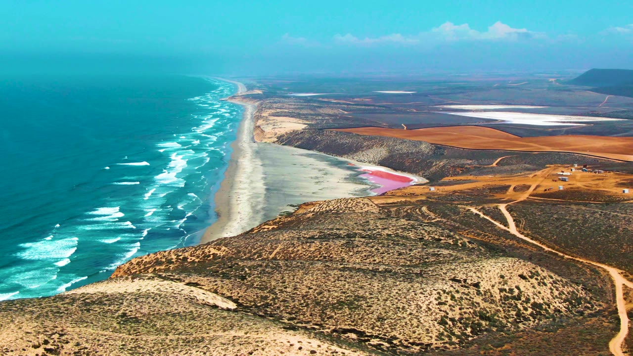 Aerial frame from San Quintin BC Mexico showing surf bands along the beach, dry hills and dirt roads in the foreground, bright inland flat areas in the distance, and no visible people.