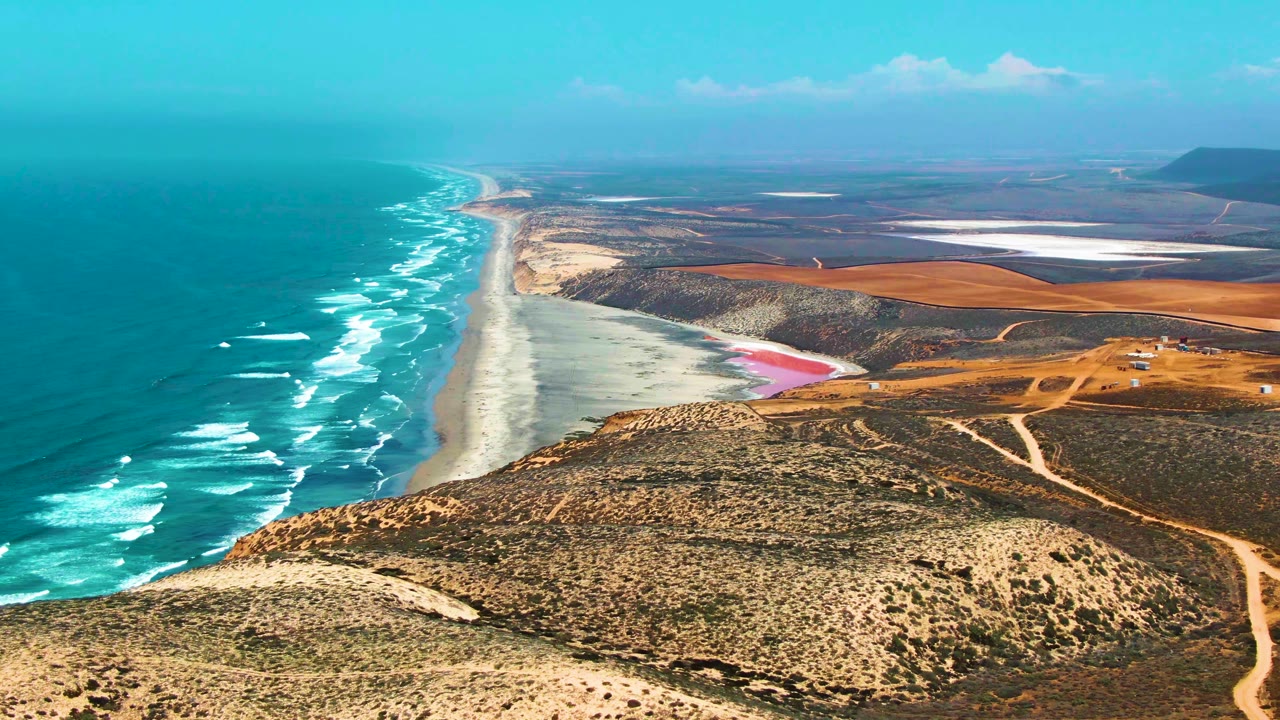 Wide aerial frame over San Quintin BC Mexico with repeating white wave lines, a narrow sandy beach, scrub hills in the foreground, reddish dry land inland, pale flats near the horizon, and no visible people.