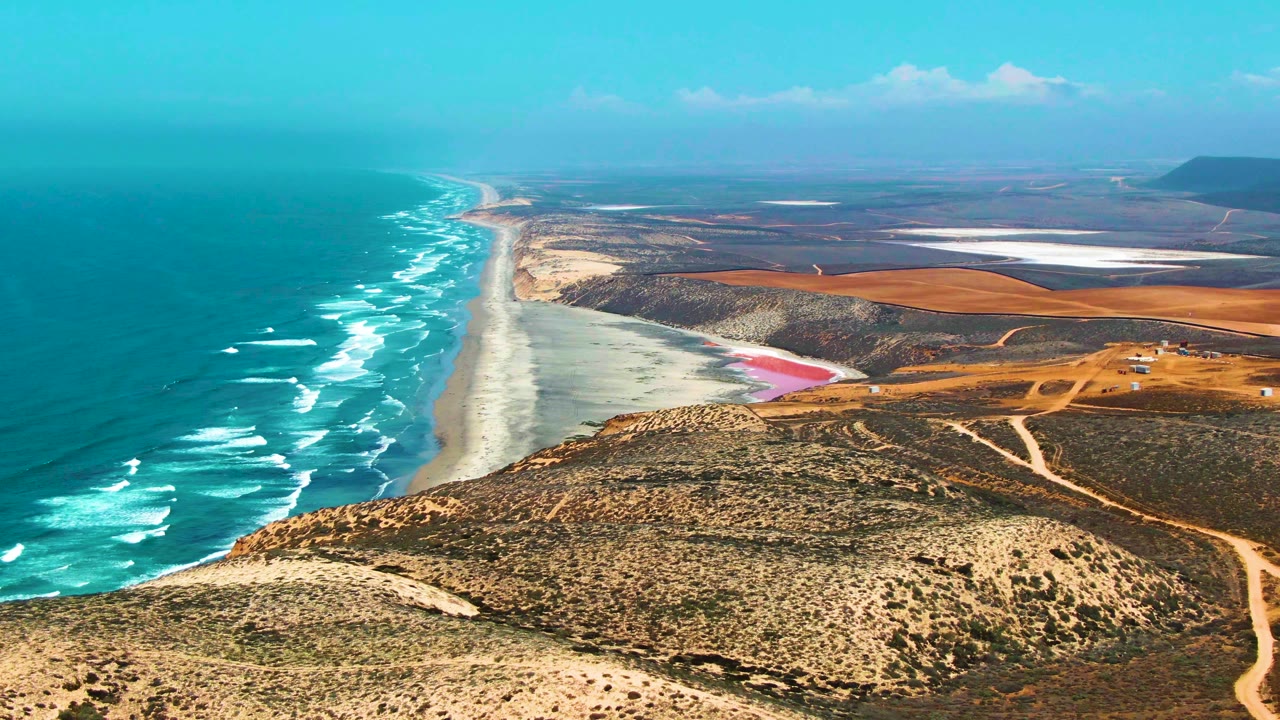 Mid-clip aerial frame in San Quintin BC Mexico, with a long diagonal coastline, white-capped waves, tan scrub hills, visible dirt tracks, bright inland flats, and no visible people.