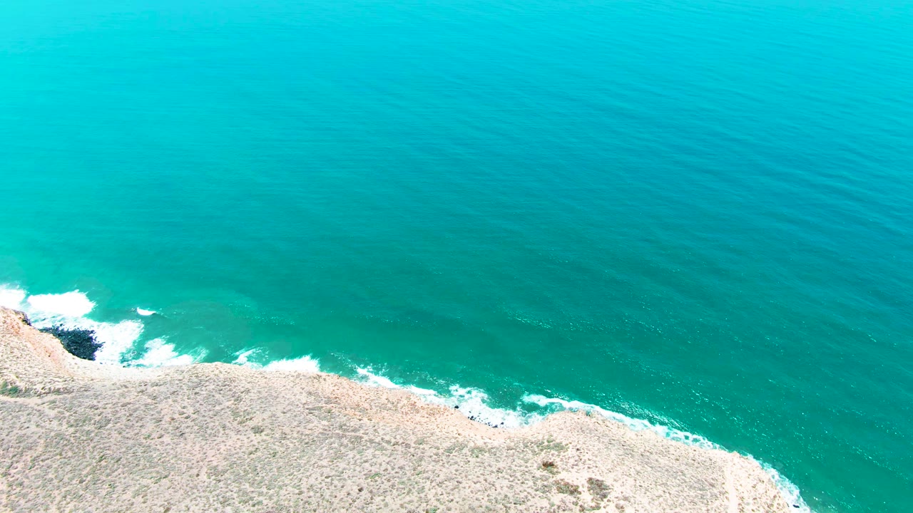 Overhead view of turquoise water meeting the pale rocky coast in San Quintin BC, Mexico, with white foam at the shore and no visible people.