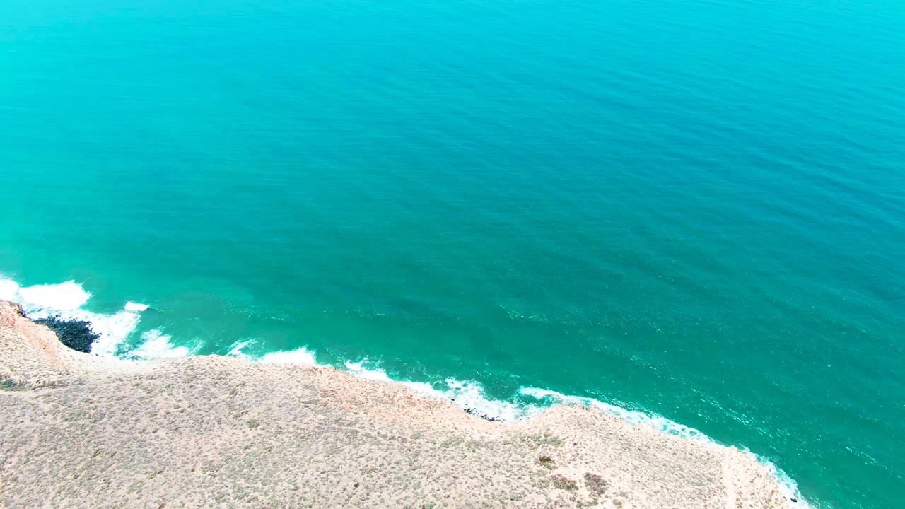 White surf curls against rocks below a broad turquoise ocean surface.