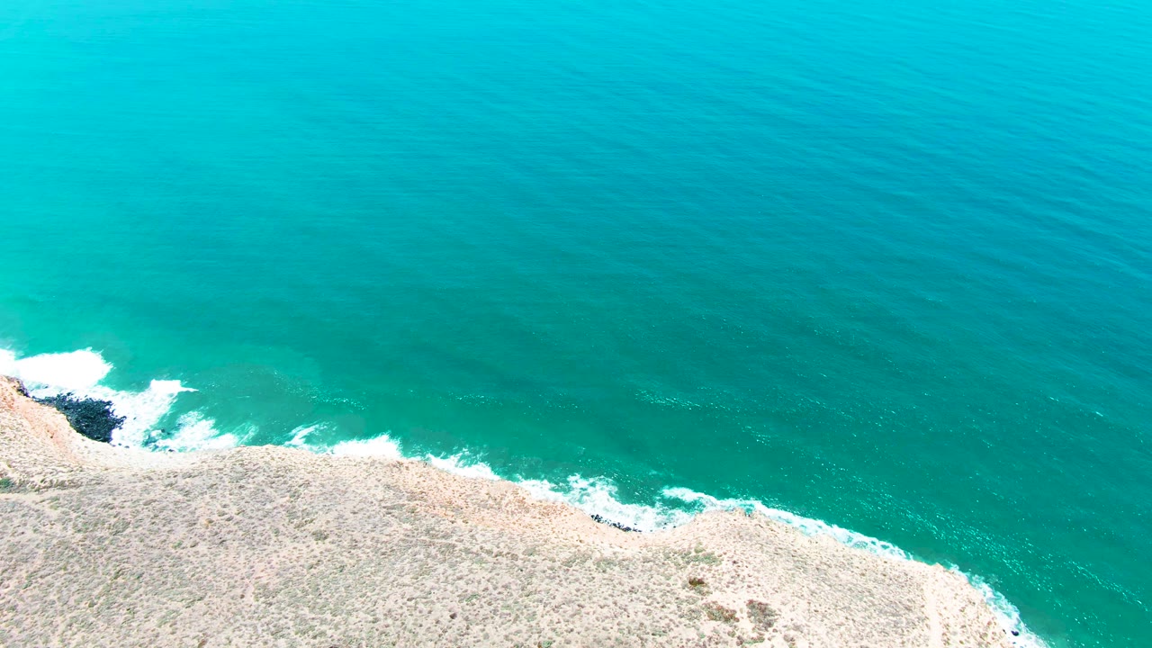 The San Quintin BC shore remains in the lower frame as fresh white foam gathers near a dark rock notch beneath the turquoise sea, with no visible people.