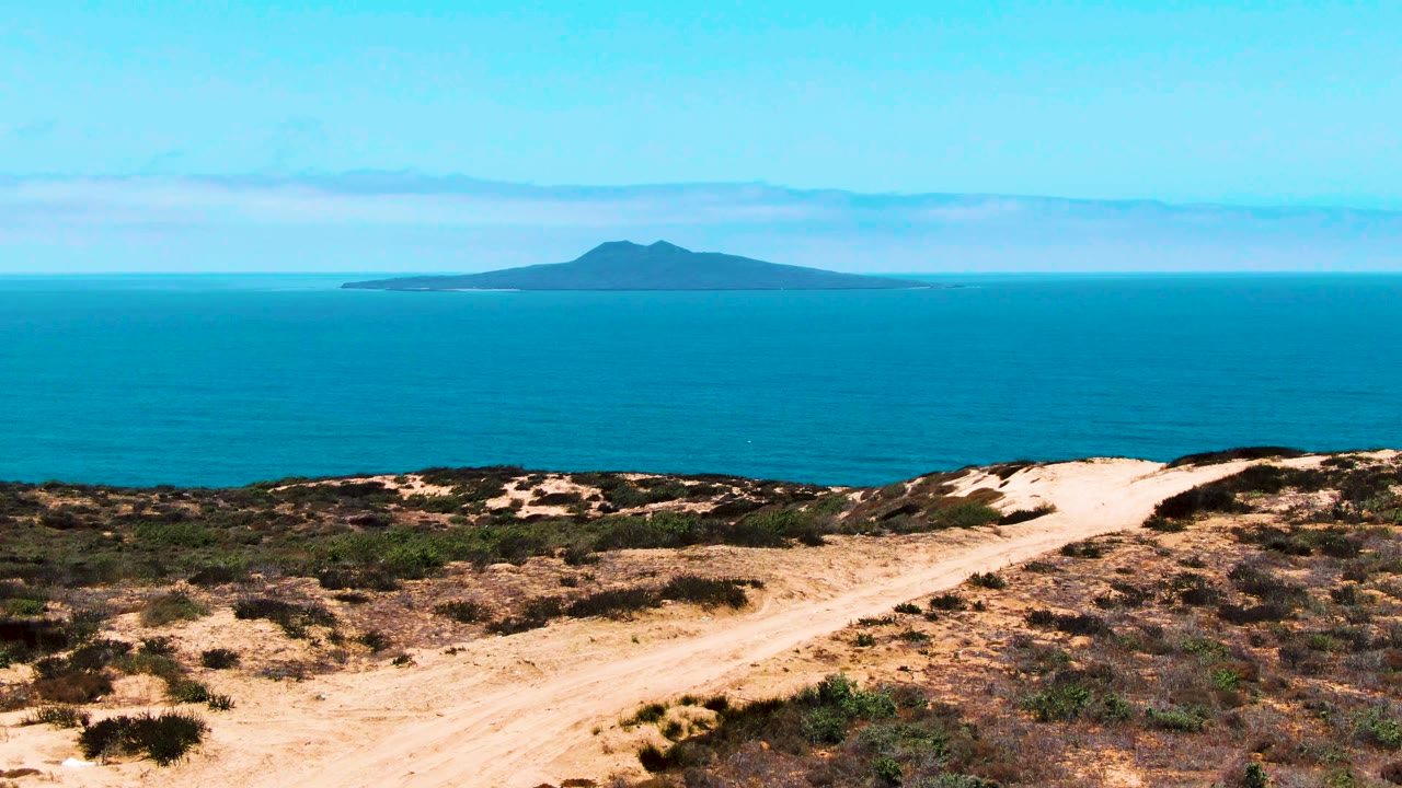 San Quintin dune track runs through sparse scrub toward a broad blue ocean plane and a distant island beneath a clear sky, with no visible people.