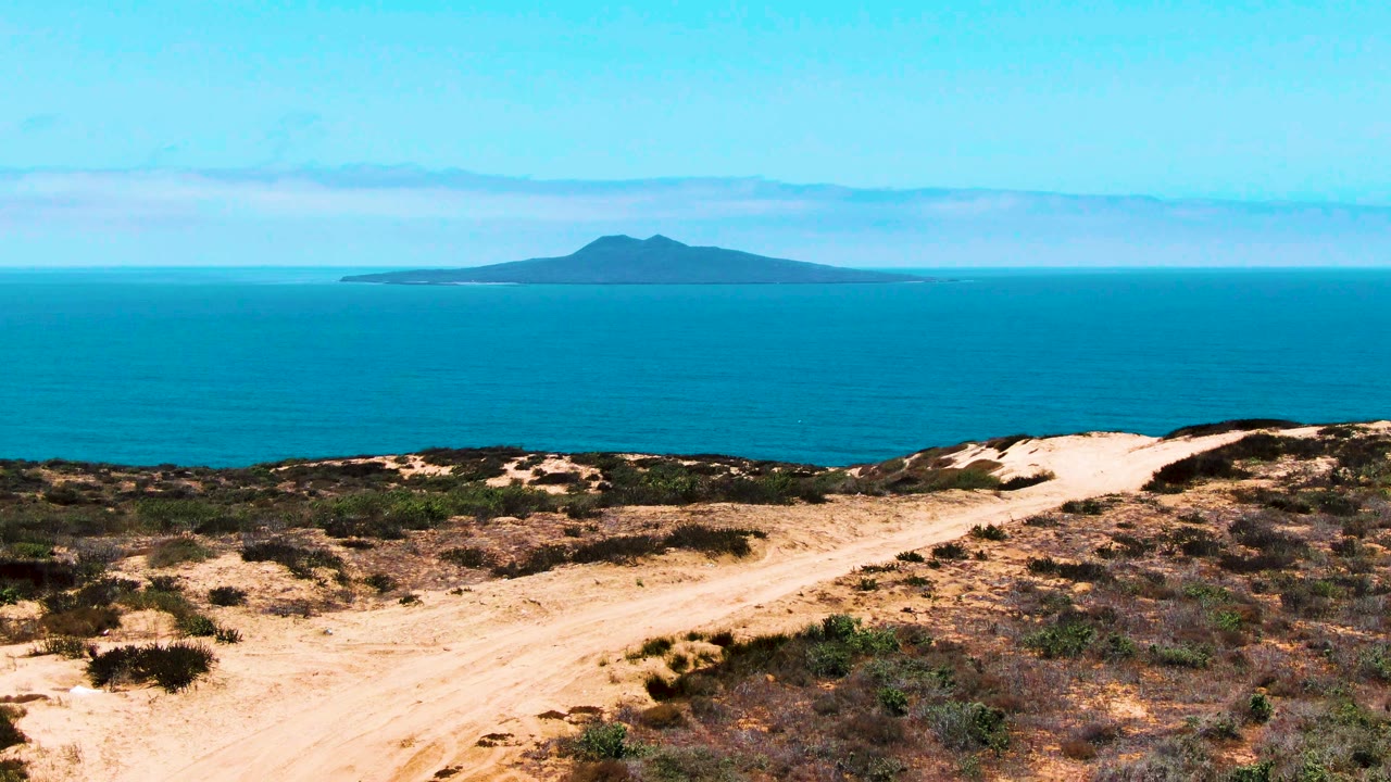 Lower-row frame shows a foreground sandy path and scrubland in San Quintin, Mexico, with calm blue water and a distant island across the horizon, and no visible people.