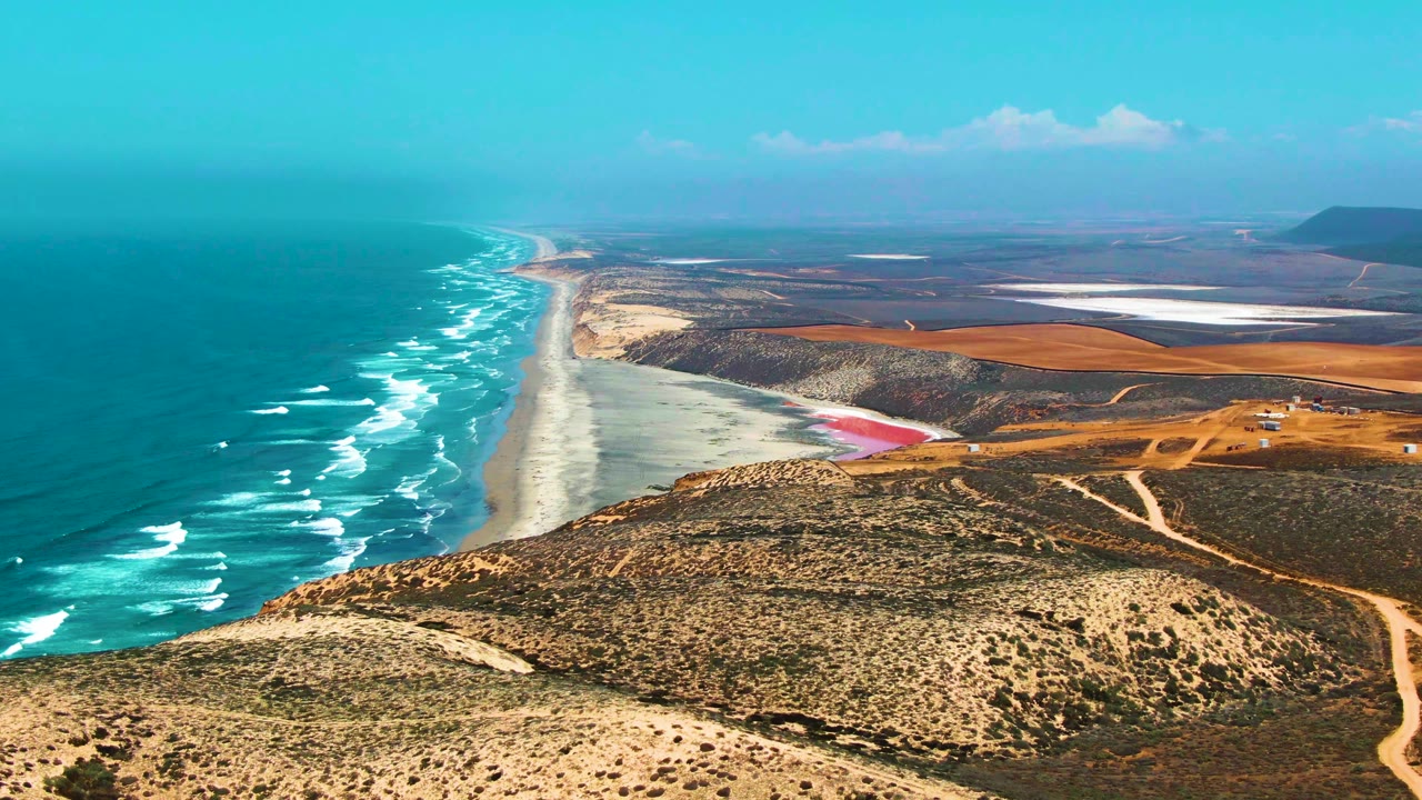 Opening aerial view over San Quintin BC Mexico coastline, with turquoise ocean and white surf on the left, scrubby hills and dirt roads in front, inland flats beyond, and no visible people.