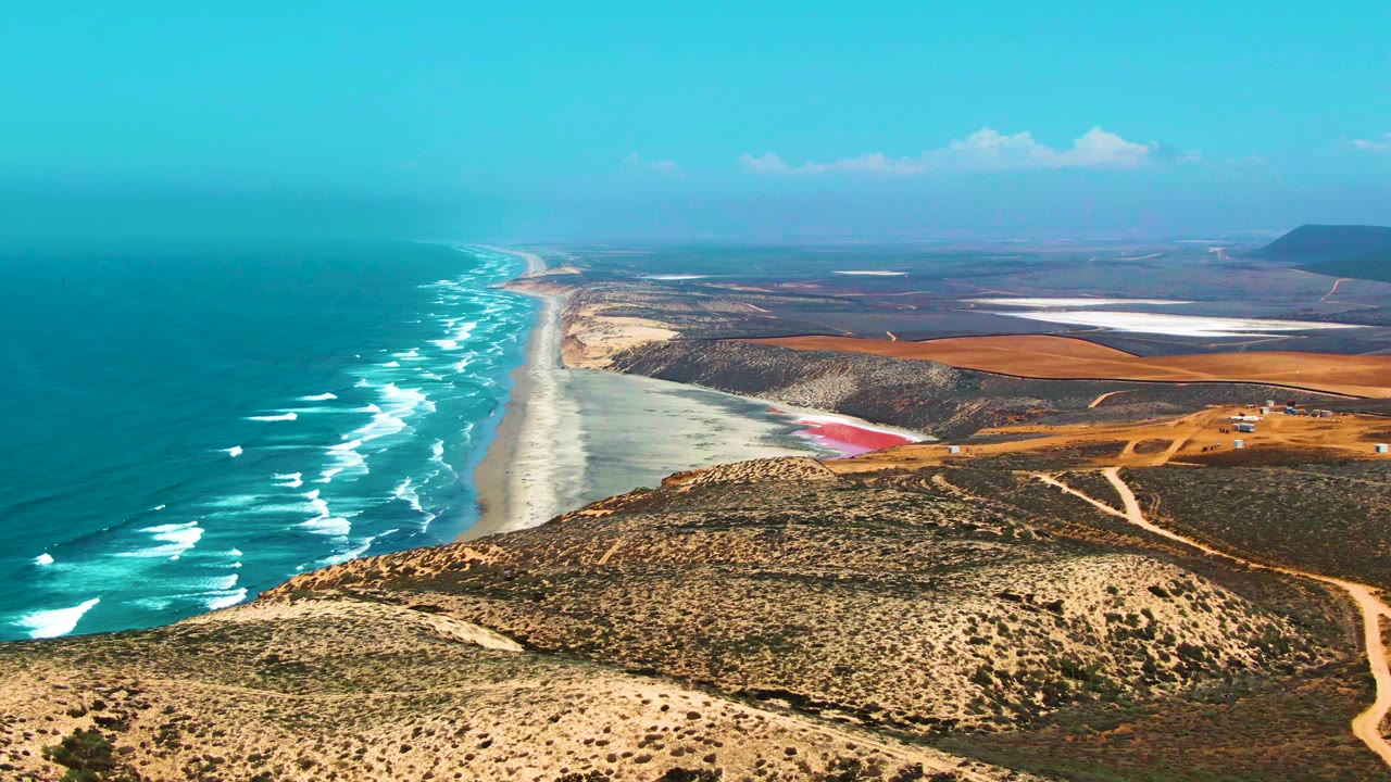 Mid-clip aerial view in San Quintin BC Mexico, with white waves advancing toward a long beach, a rounded scrubby hill in the foreground, dirt roads to the right, and no visible people.
