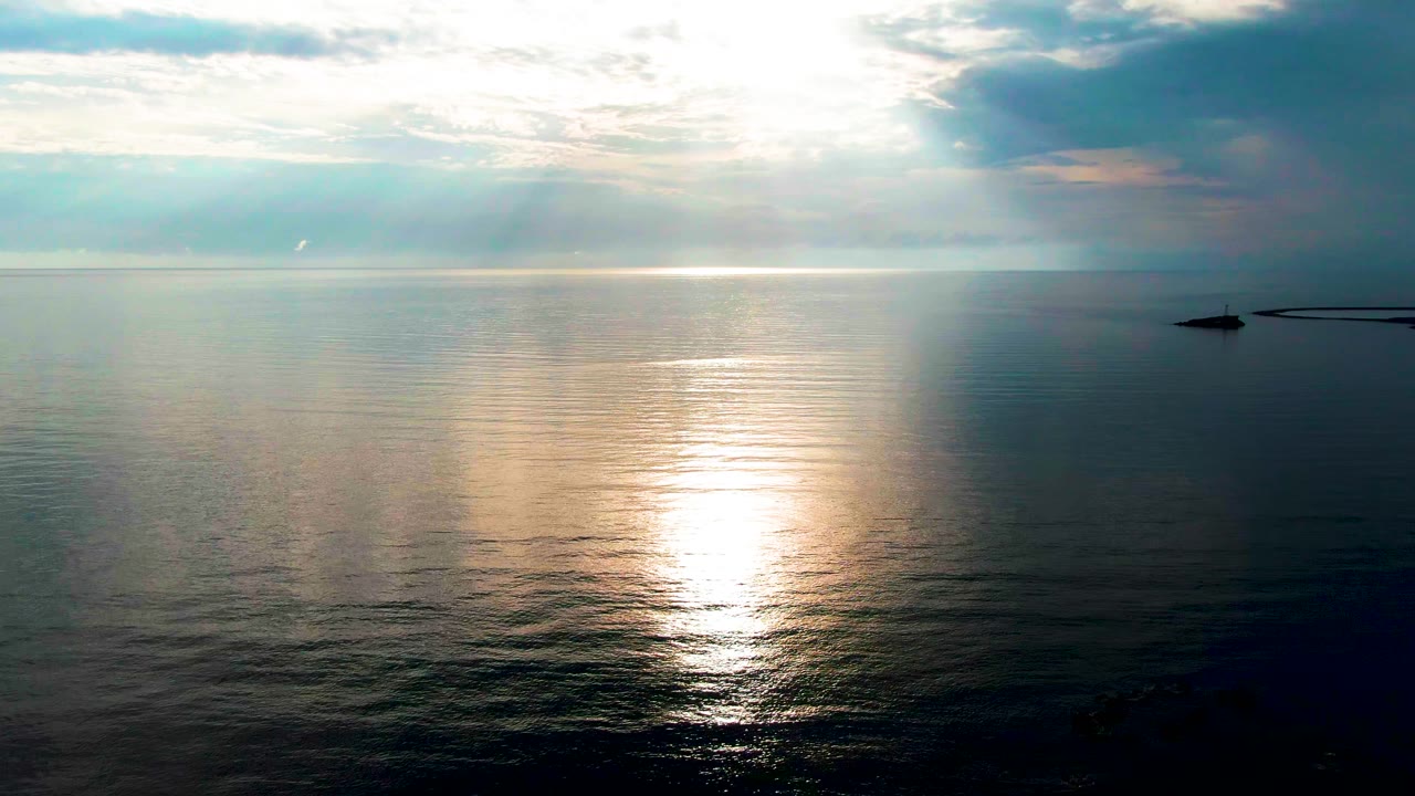 Calm ocean off Santa Rosalia, Baja California Sur reflects a bright band of sunlight under layered clouds, with a small dark point of land at far right and no visible people.