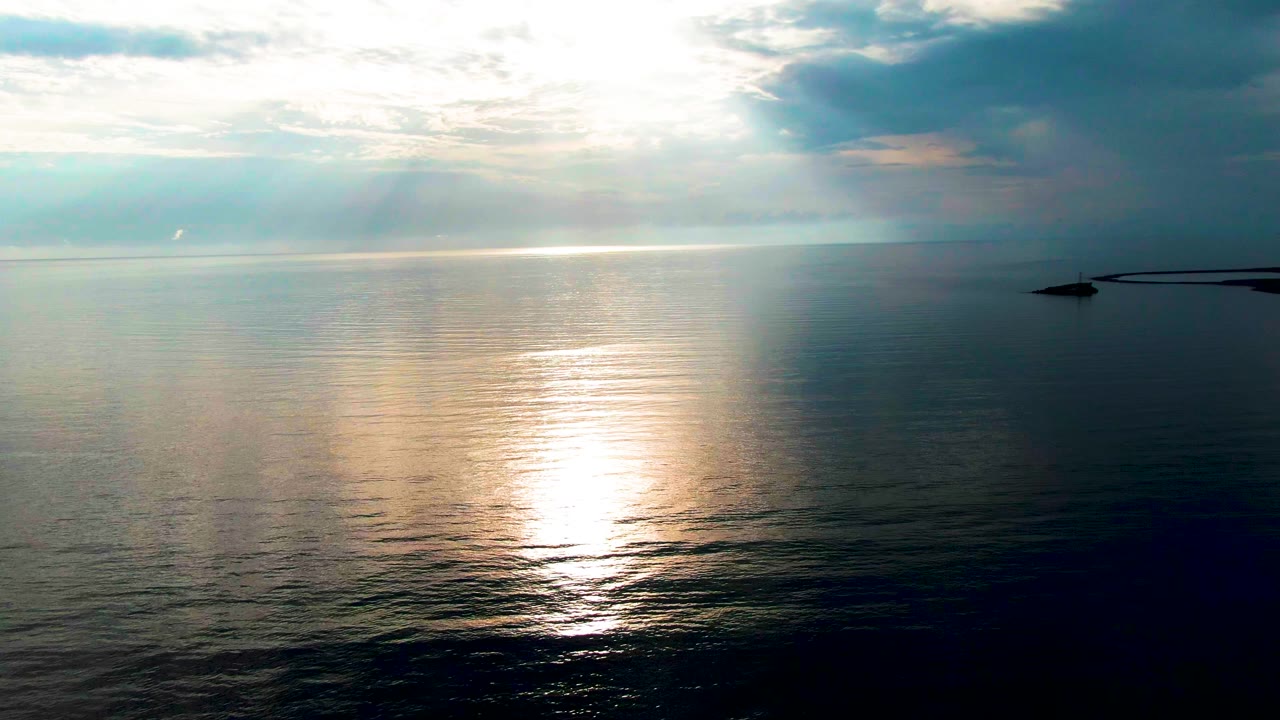 At 0:01 in Santa Rosalia, Baja California Sur, the bright water reflection sits slightly lower in the frame, with soft wave texture across the foreground and the small dark pier or jetty shape near the right edge, no visible people.
