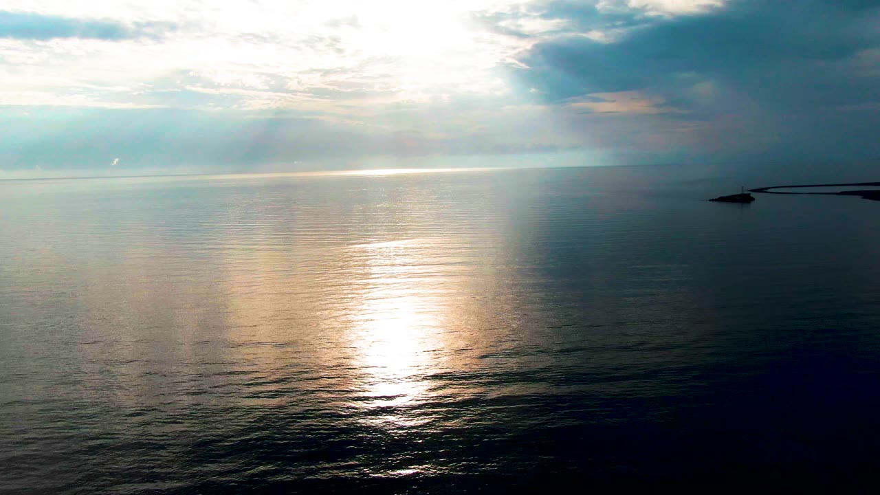 At 0:02 in Santa Rosalia, Baja California Sur, the sun reflection spreads across the mid-ocean while a darker cloud mass hangs over the right side of the horizon and the distant structure remains visible, no visible people.