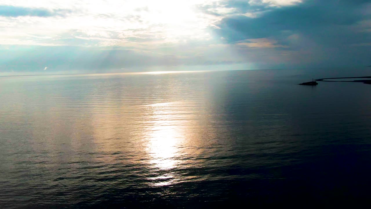 At 0:03 in Santa Rosalia, Baja California Sur, a wide high view holds on the ocean surface as the reflection breaks into smaller highlights across the ripples, with the right-side structure partly silhouetted, no visible people.