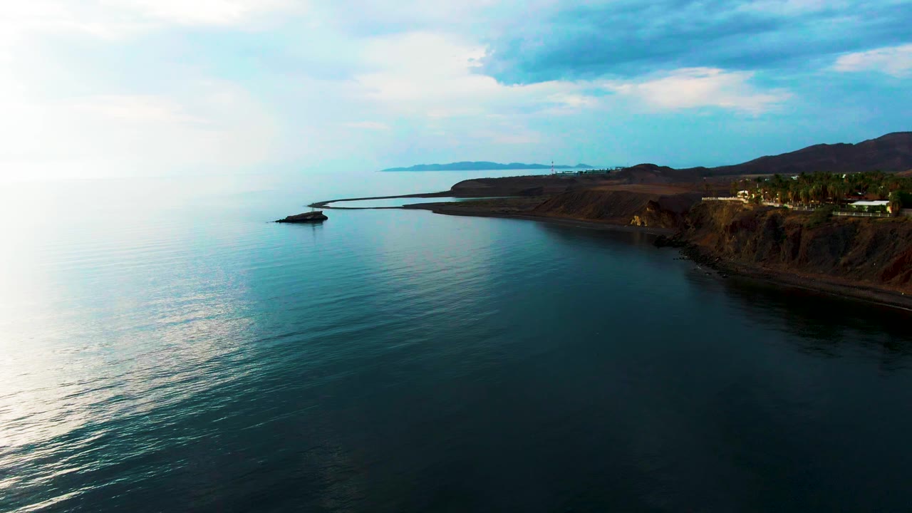 At 0:02, reflective water occupies the foreground while the Santa Rosalia cliffline, small buildings, and low hills remain along the right side with no visible people.