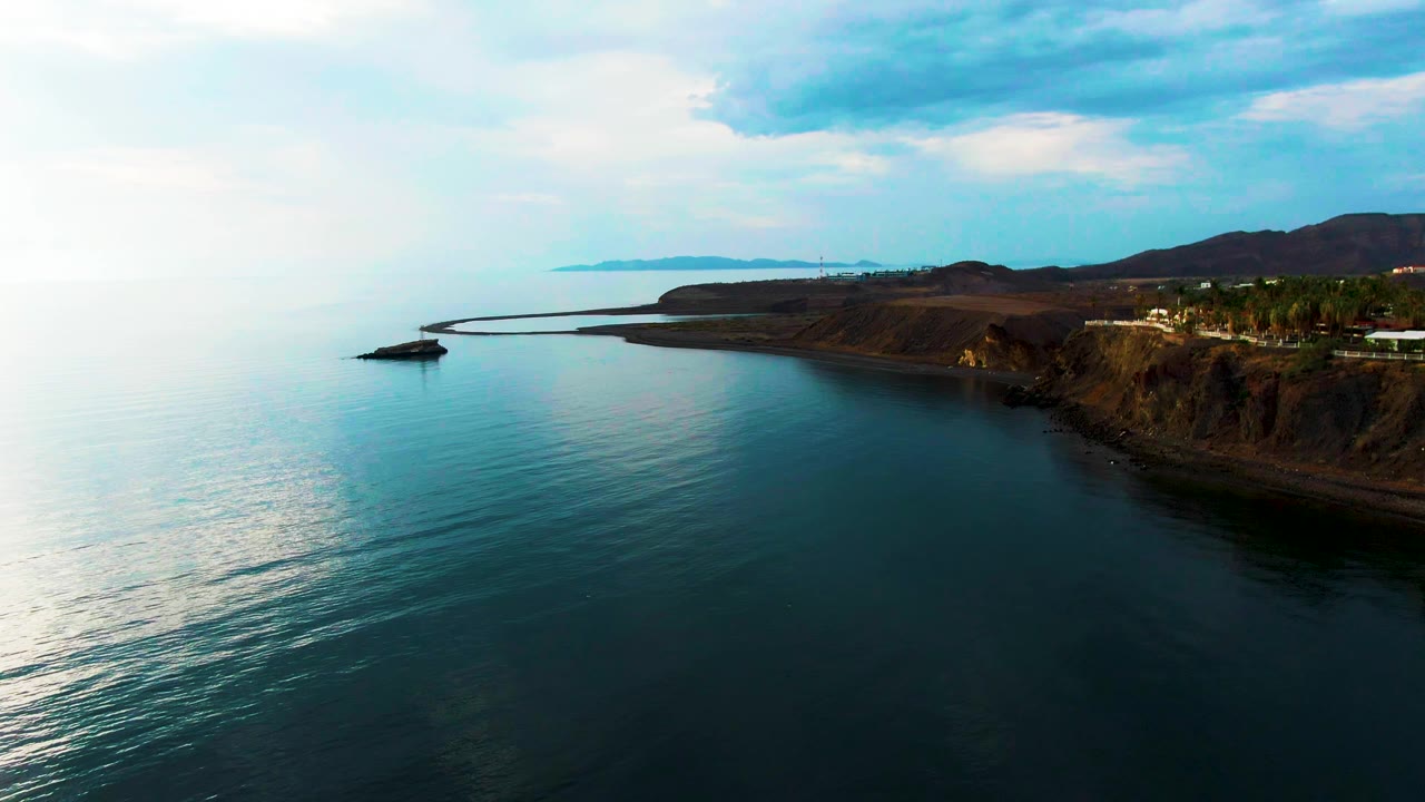 Aerial frame over Santa Rosalia, Baja California Sur, with calm blue ocean in the foreground, rocky cliffs on the right, a curved shoreline ahead, distant land on the horizon, and no visible people.