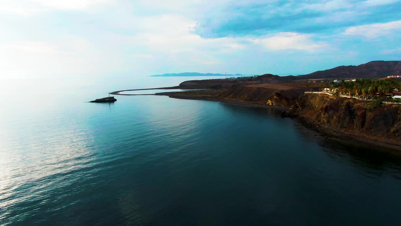 The Santa Rosalia shoreline remains in a wide aerial composition, with calm ocean below, a curved breakwater extending from the coast, cliffs on the right, distant hills, and no visible people.