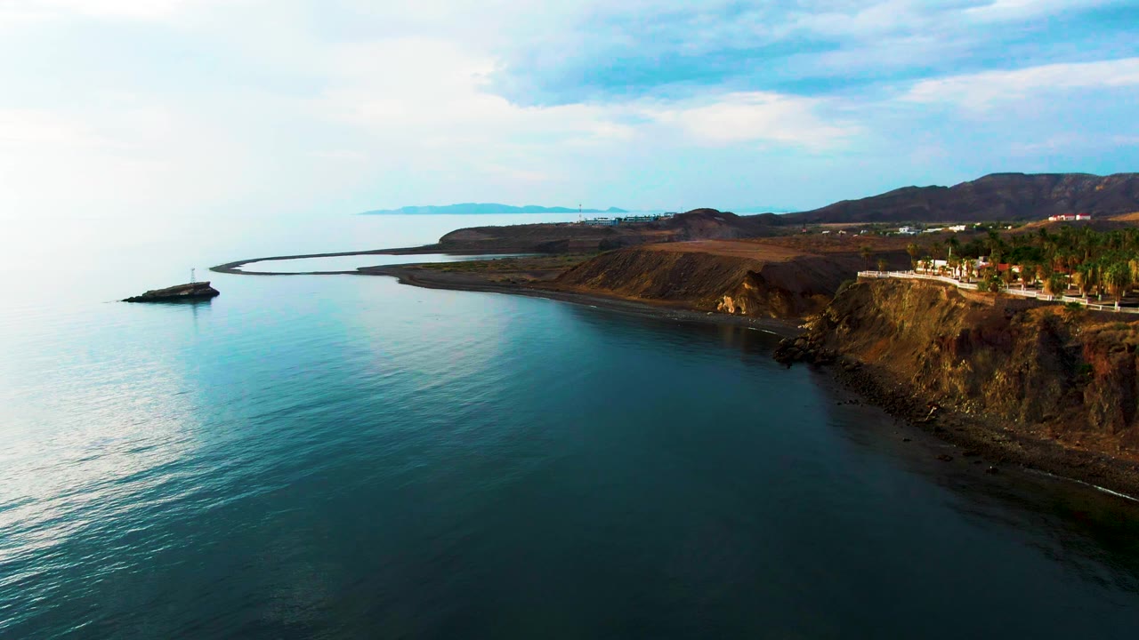 Aerial view in Santa Rosalia showing glassy blue ocean, a curved coastal edge, a road and sparse trees on the cliff, distant hills, and no visible people.