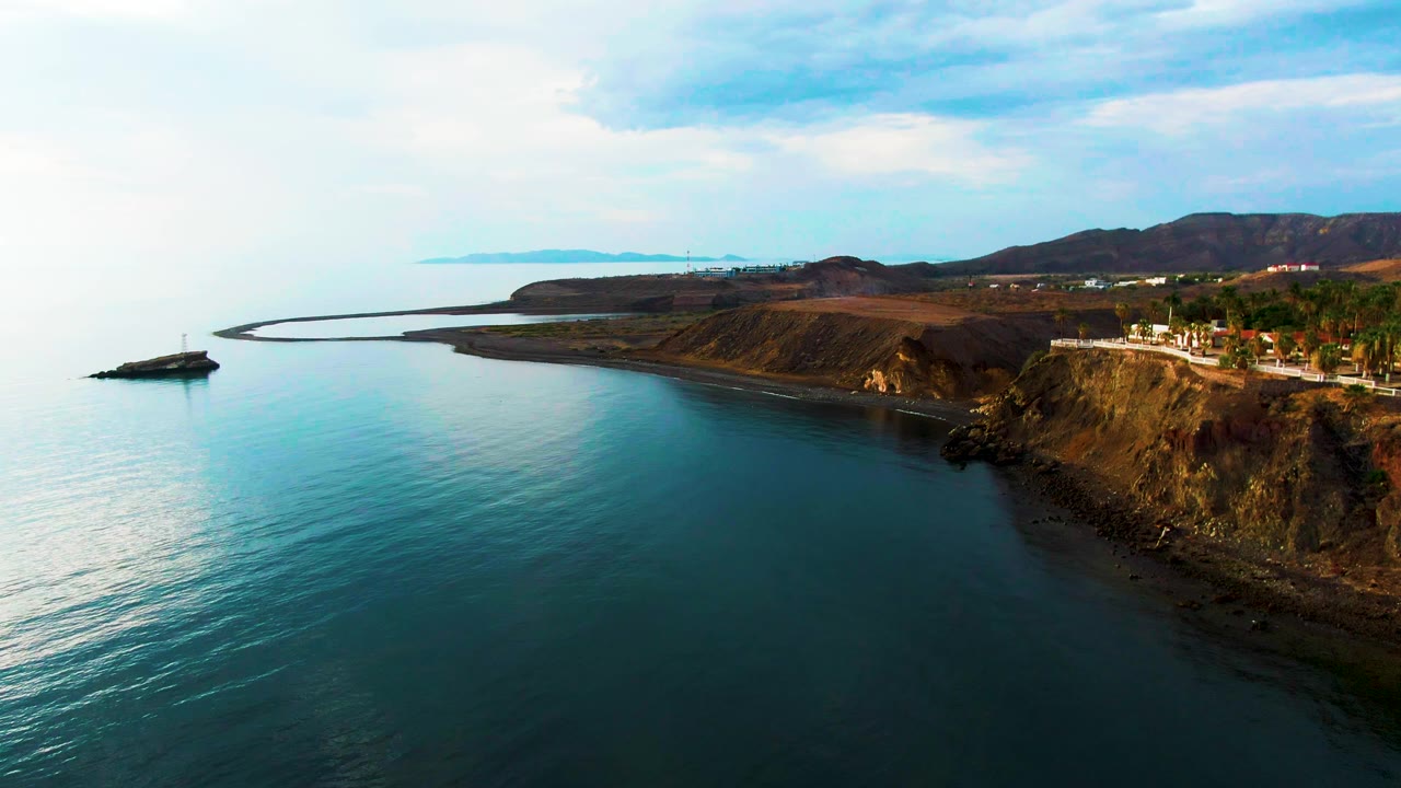 Mid-clip aerial frame over Santa Rosalia with calm teal water below, the cliff road more centered along the right edge, dry hills behind it, and no visible people.