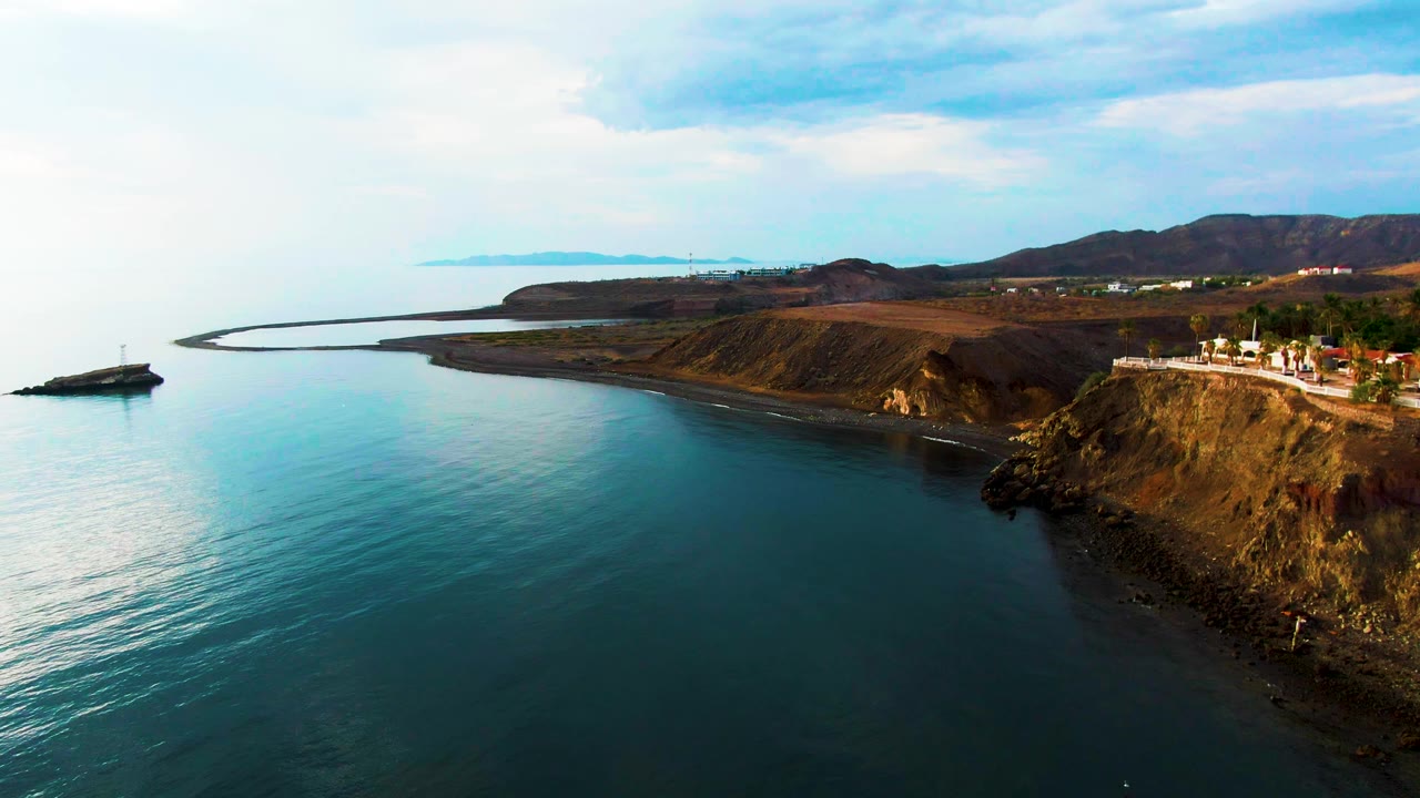 Coastal road and brown cliffs above calm blue sea under a partly cloudy sky.