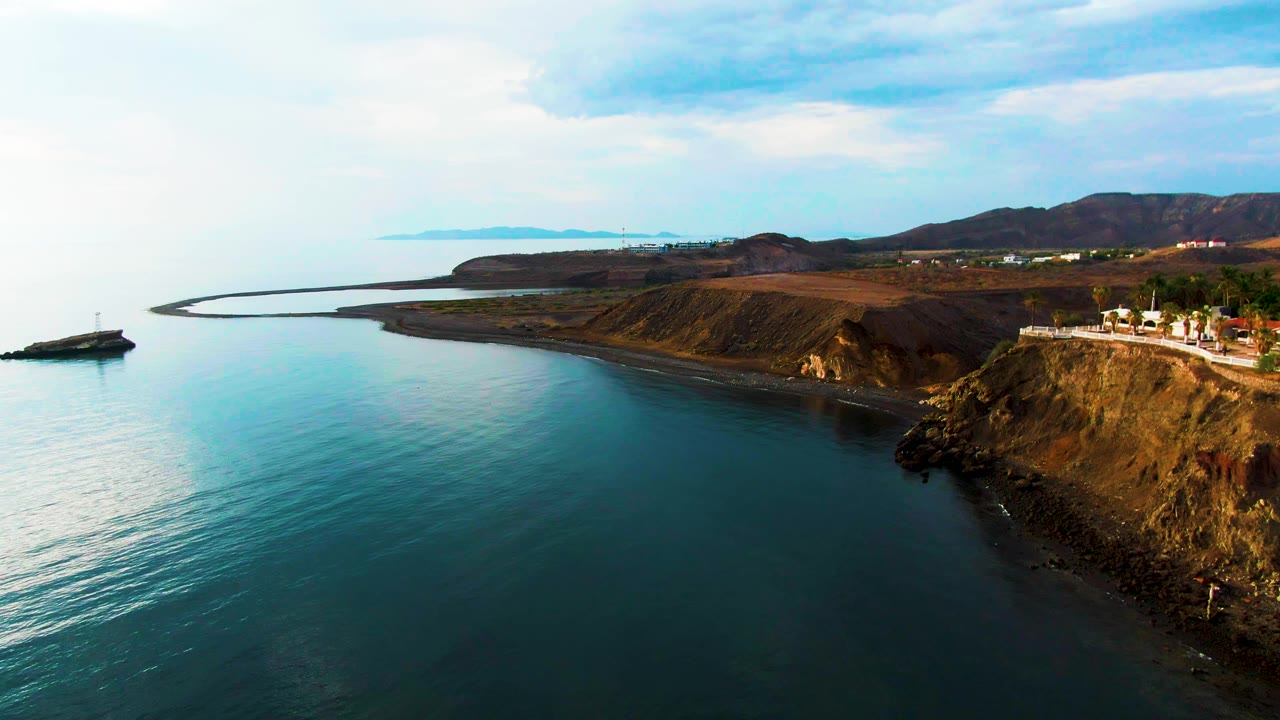 Santa Rosalia coastline from an elevated drone angle, with the cliff road crossing the right side, smooth water below, low hills beyond, and no visible people.