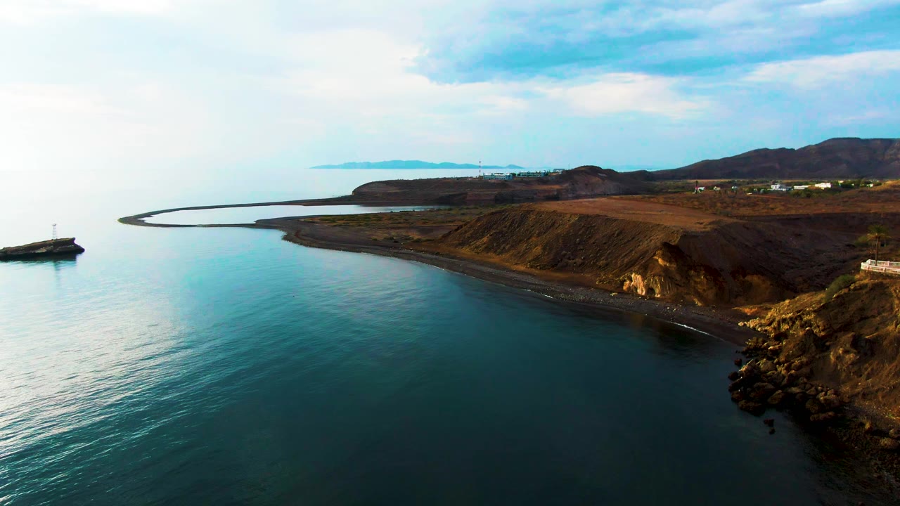 Aerial view over Santa Rosalia, Baja California Sur, with calm blue-green water, a dark rocky shoreline, low hills, a curved harbor wall, and no visible people.